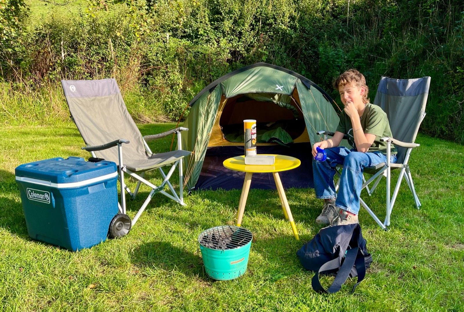 A boy sits in a camping chair beside a small green tent in a grassy field, with a cool box, a yellow table and a bucket barbecue