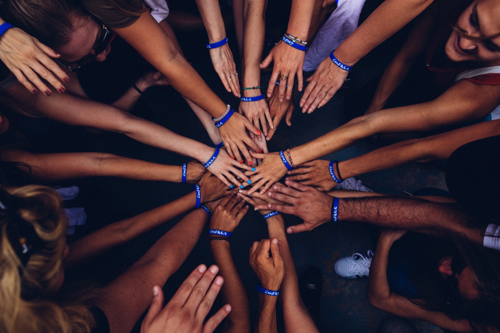 Seen from above, a circle of people reach in and stack their hands together in the middle, most wearing matching blue wristbands